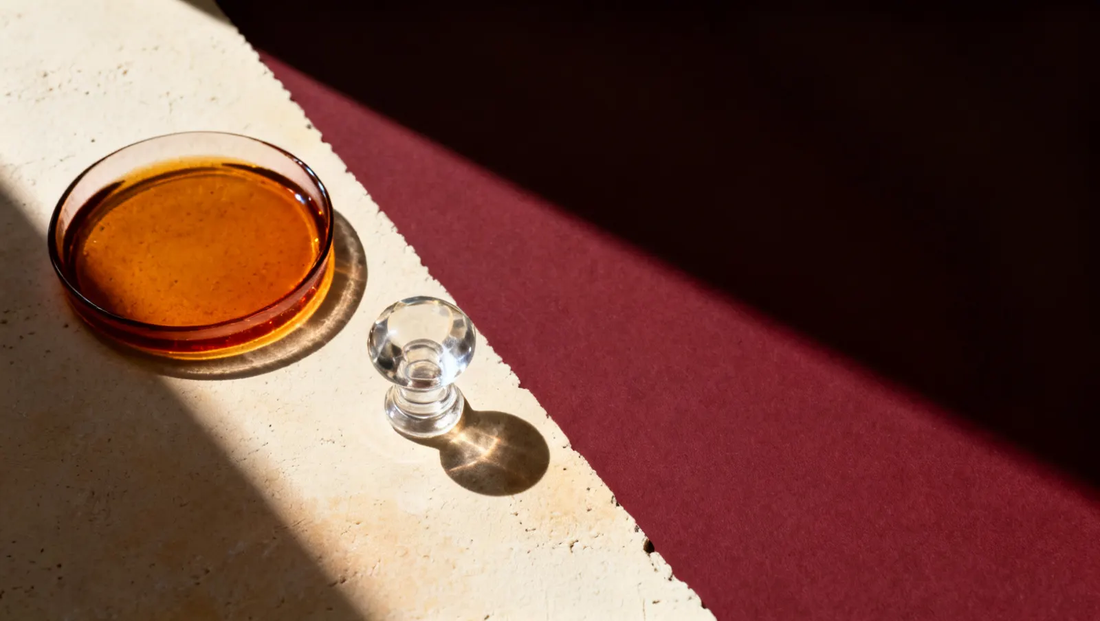 Natureza-morta em luz lateral dura: um prato raso com liquido ambar e uma tampa pesada de vidro transparente, sobre fundo vinho e creme