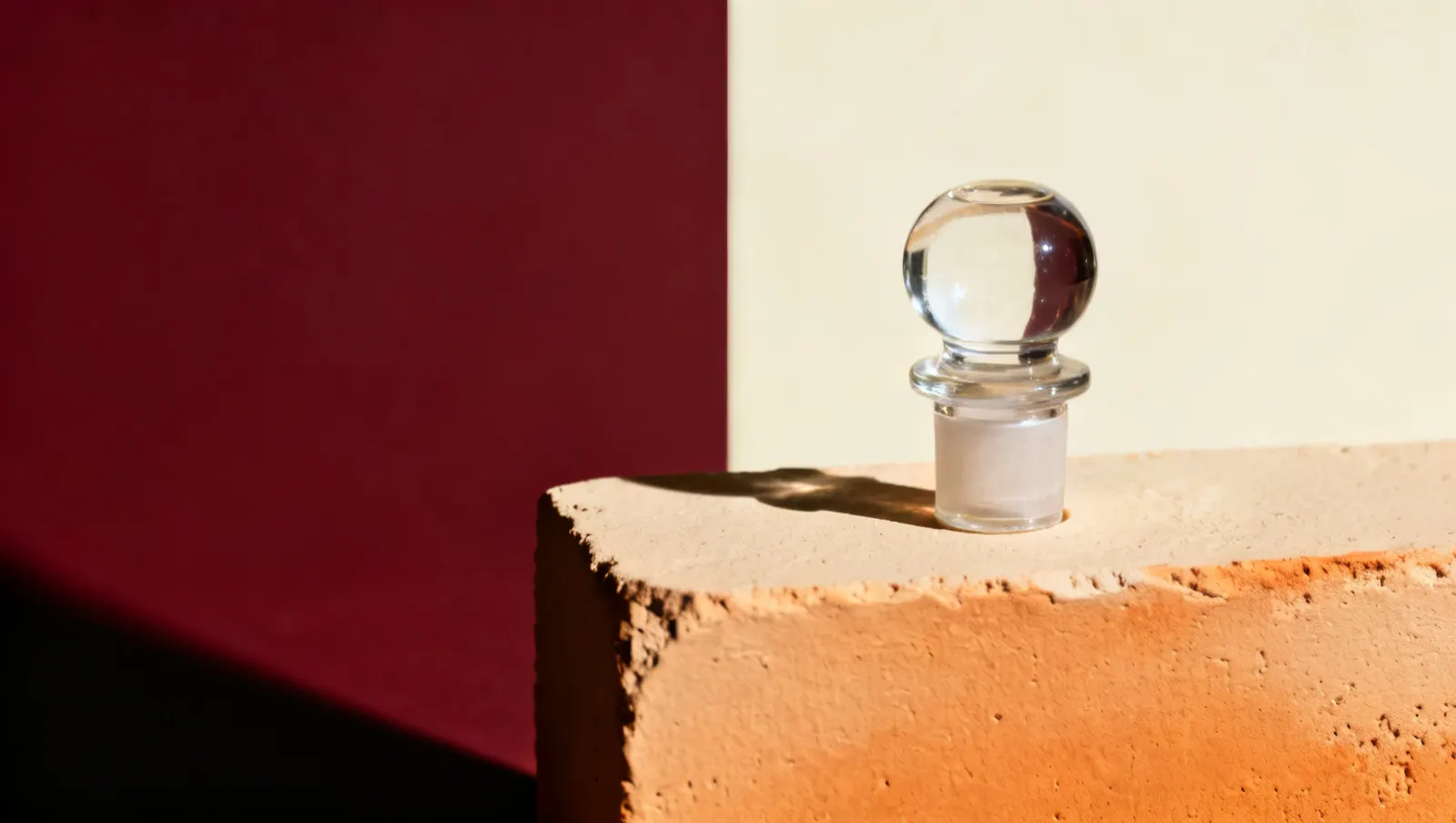 Natureza-morta em luz lateral dura: uma tampa pesada de vidro transparente, sobre fundo vinho e creme