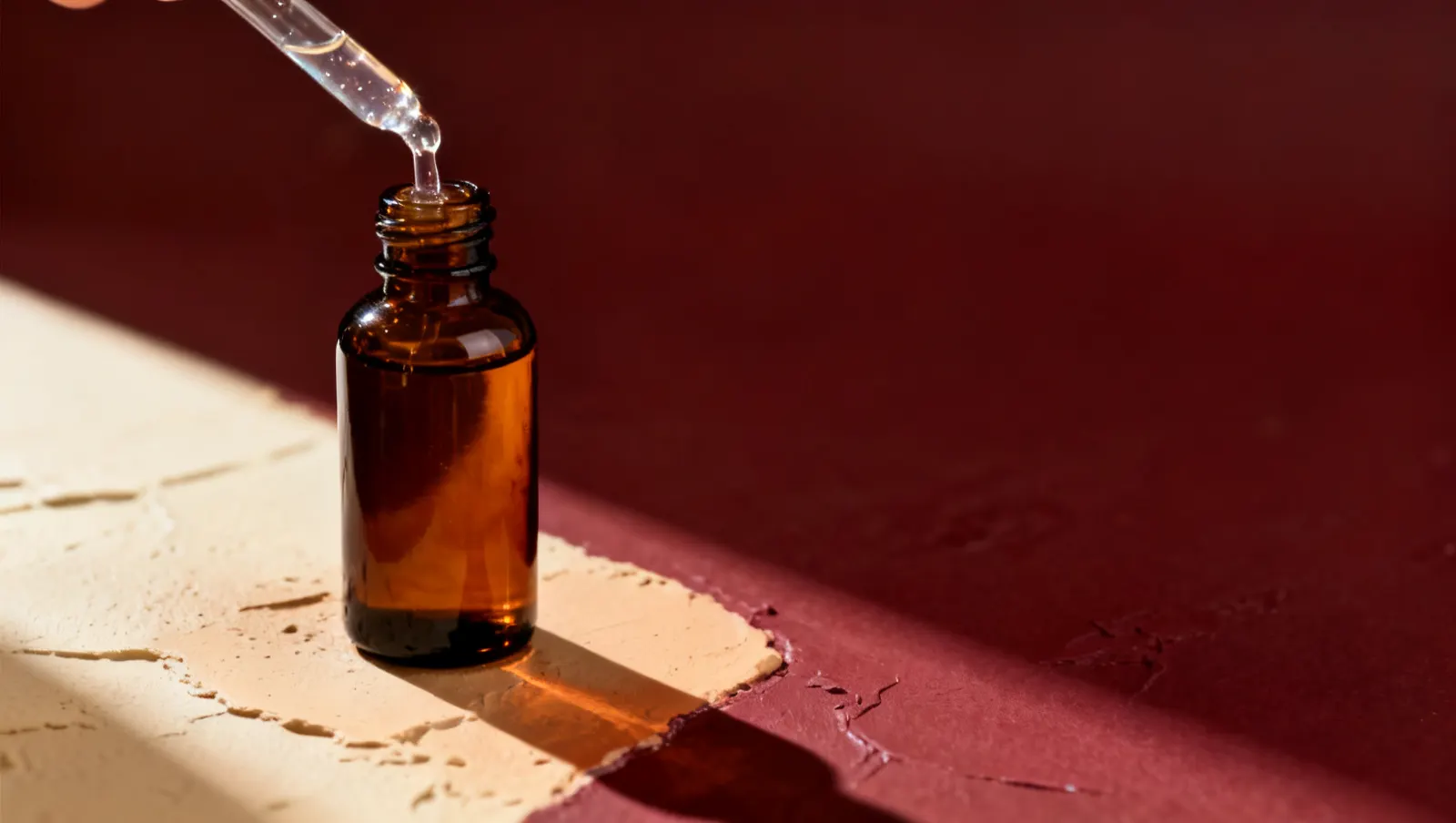 Natureza-morta em luz lateral dura: um fio de gel transparente espesso e um frasco de vidro ambar sem rotulo, sobre fundo vinho e creme