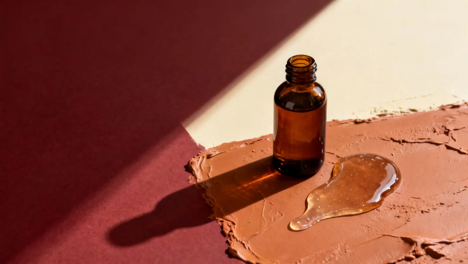 Natureza-morta em luz lateral dura: um frasco de vidro ambar sem rotulo e um fio de gel transparente espesso, sobre fundo vinho e creme