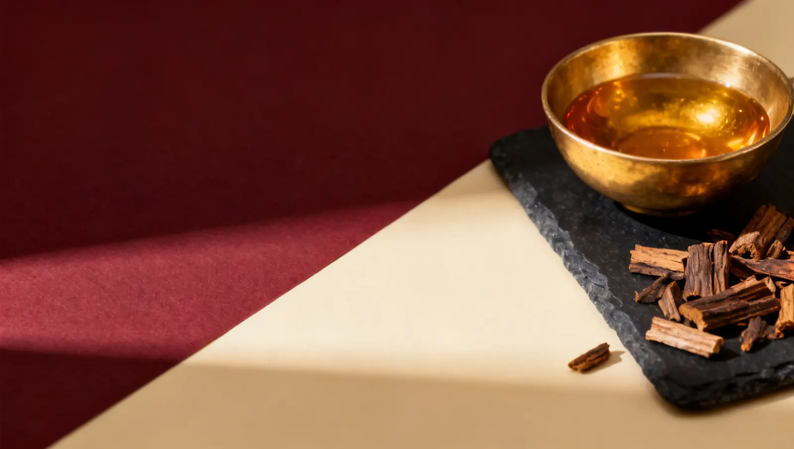 Natureza-morta em luz lateral dura: lascas de madeira de oud e uma tigela lisa de latao, sobre fundo vinho e creme