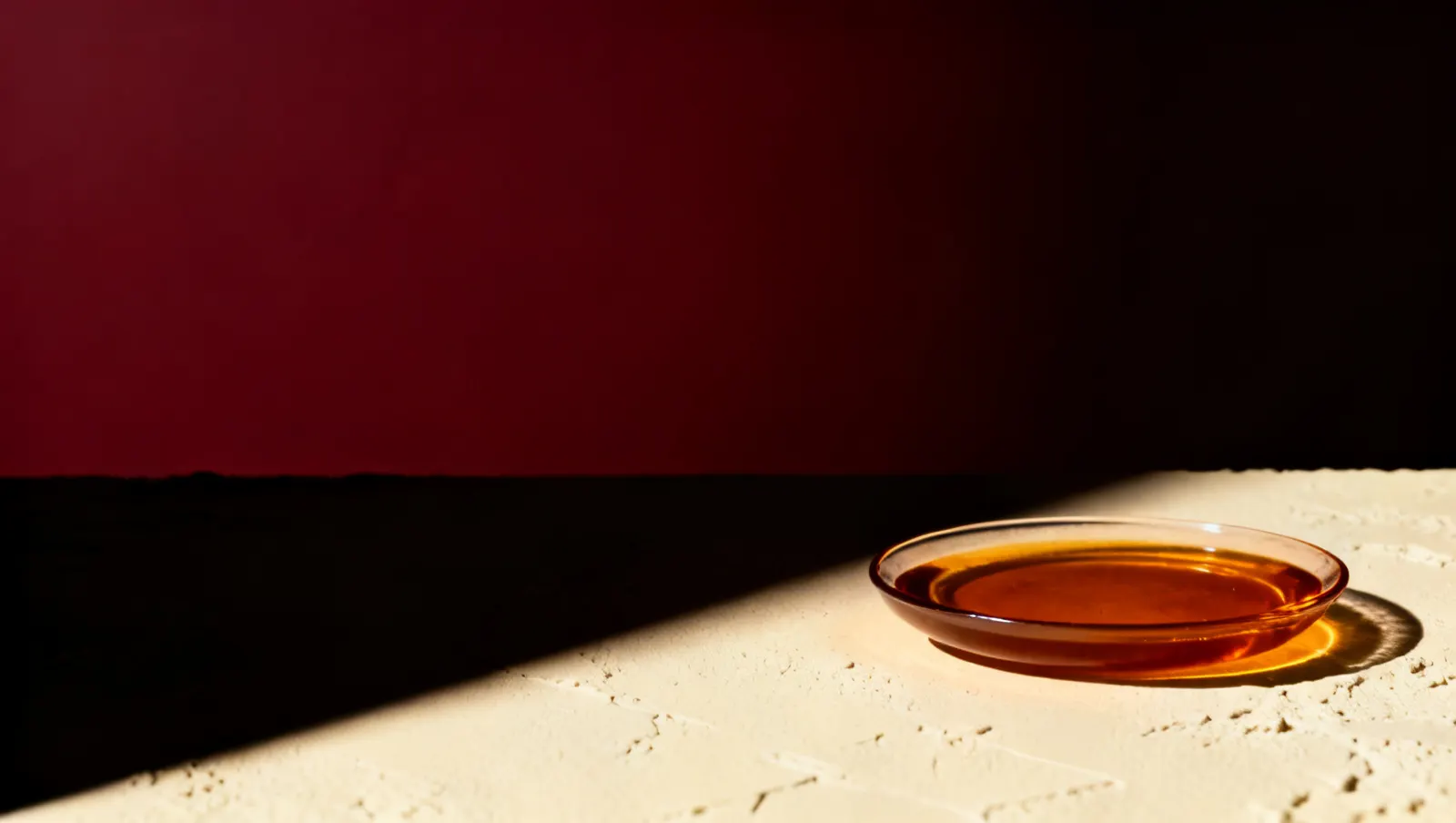 Natureza-morta em luz lateral dura: um prato raso com liquido ambar, sobre fundo vinho e creme