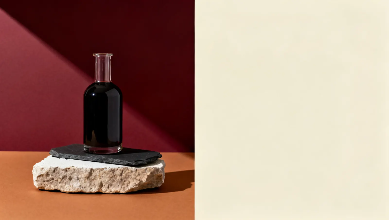 Natureza-morta em luz lateral dura: uma placa de pedra bruta e um recipiente pesado de vidro com liquido escuro, sobre fundo vinho e creme