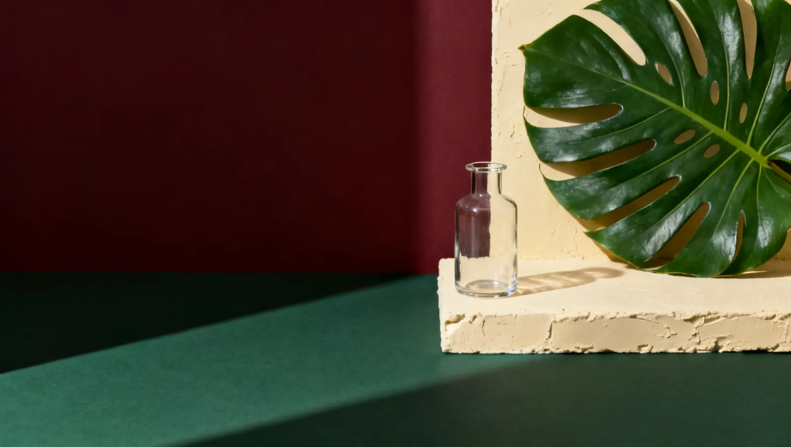Natureza-morta em luz lateral dura: um recipiente de vidro sem rotulo e uma folha tropical grande, sobre fundo vinho e creme