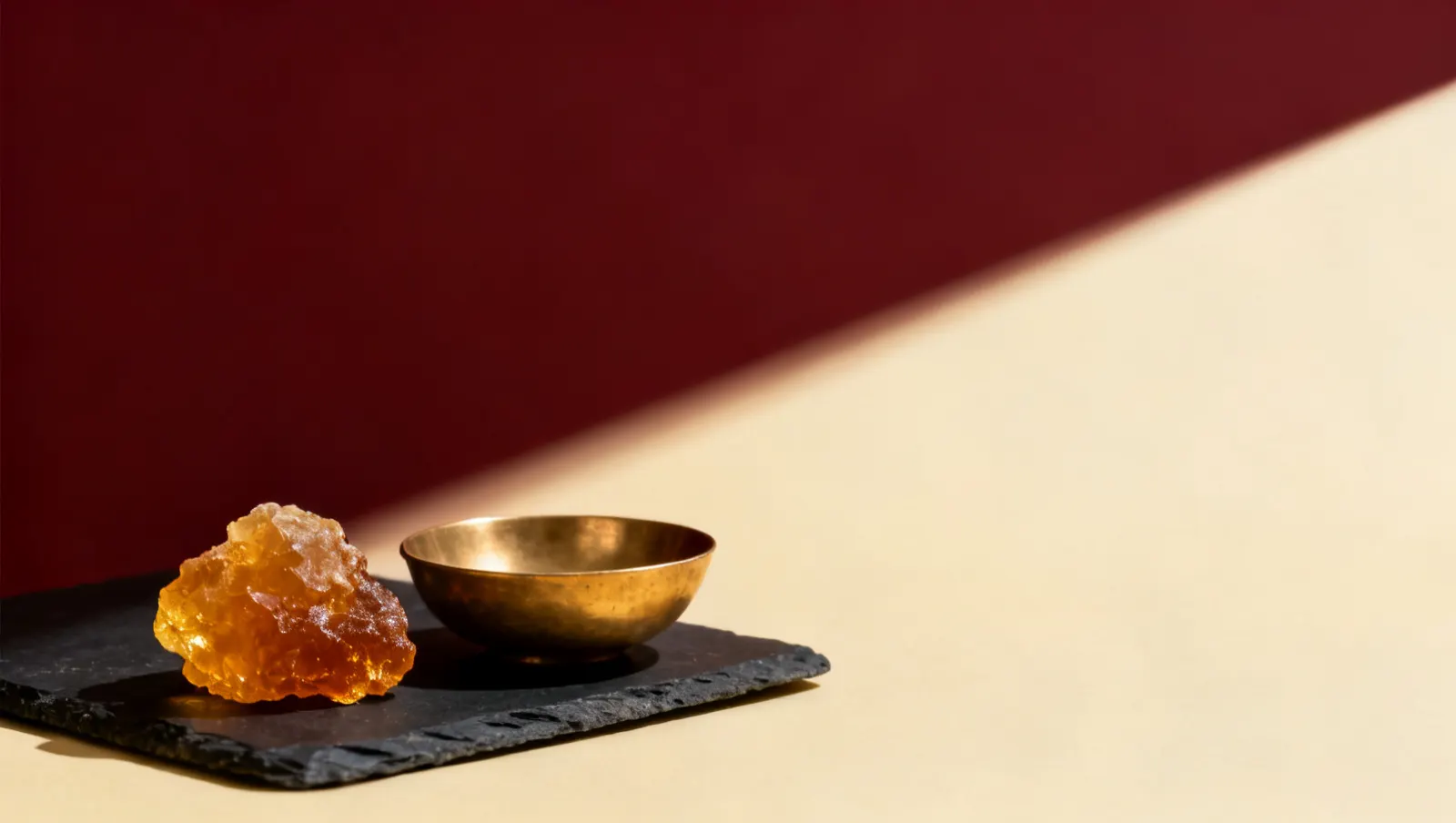 Natureza-morta em luz lateral dura: um bloco de resina de ambar dourado e uma tigela lisa de latao, sobre fundo vinho e creme