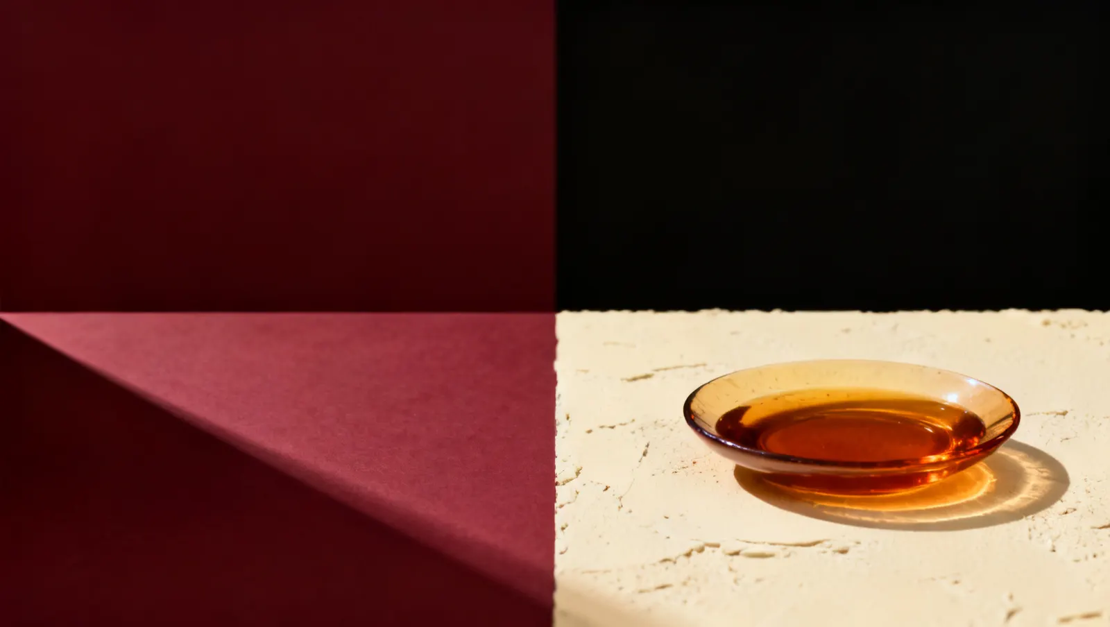 Natureza-morta em luz lateral dura: um prato raso com liquido ambar, sobre fundo vinho e creme