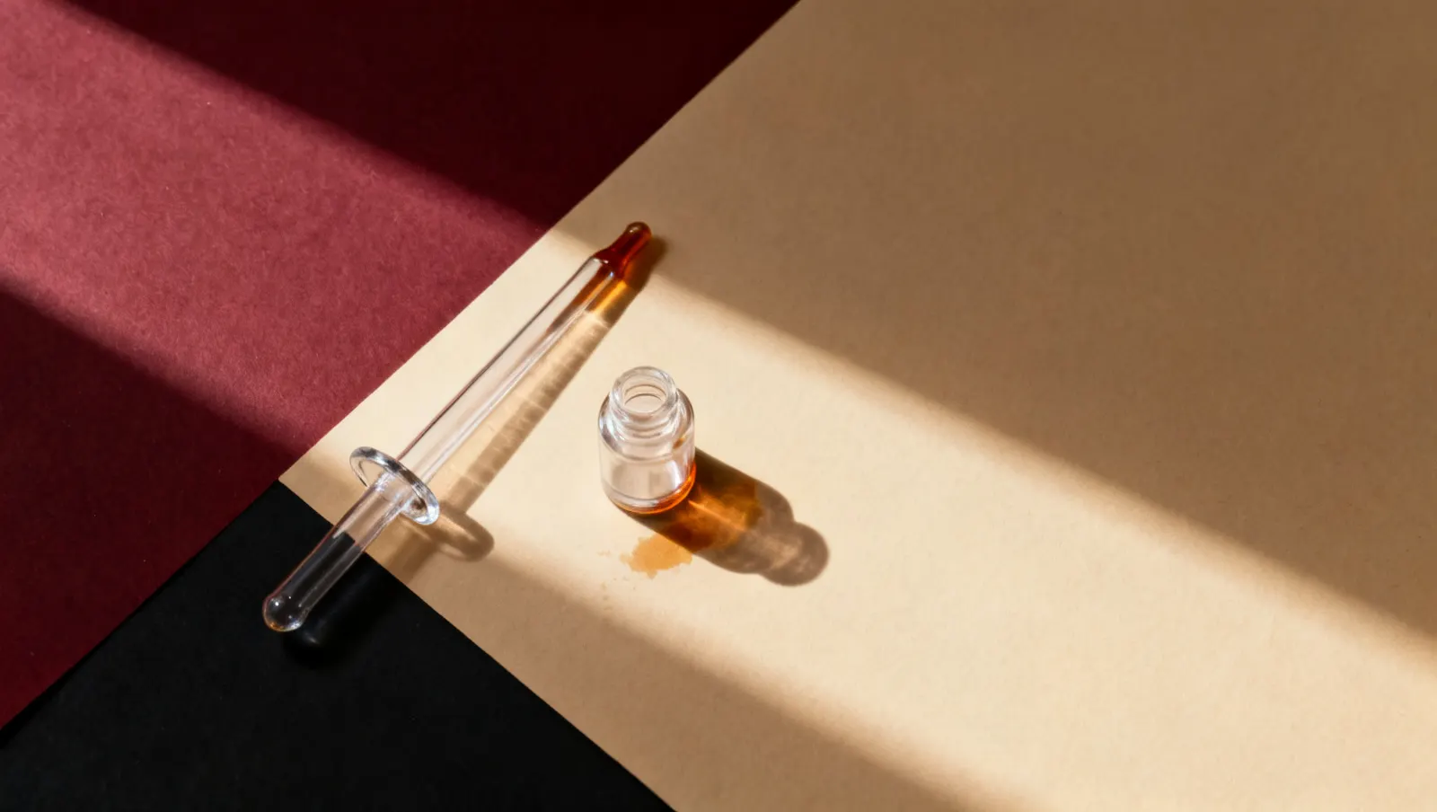 Natureza-morta em luz lateral dura: uma pipeta de vidro lisa e um frasco pequeno de vidro transparente, sobre fundo vinho e creme