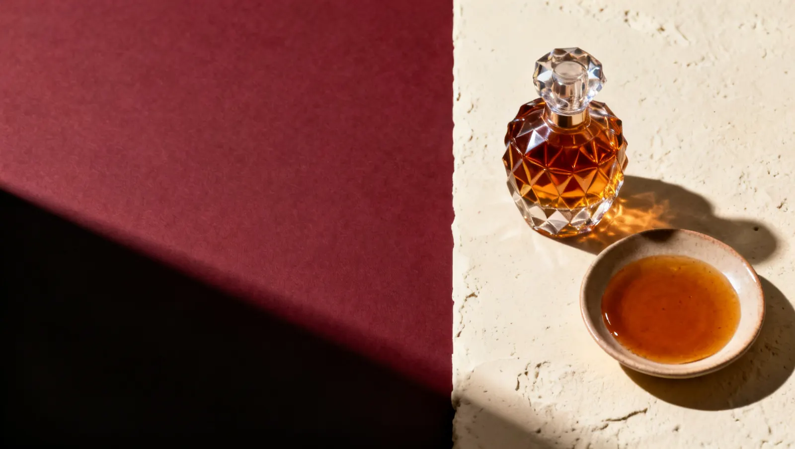 Natureza-morta em luz lateral dura: um frasco de cristal facetado sem rotulo e um prato raso com liquido ambar, sobre fundo vinho e creme
