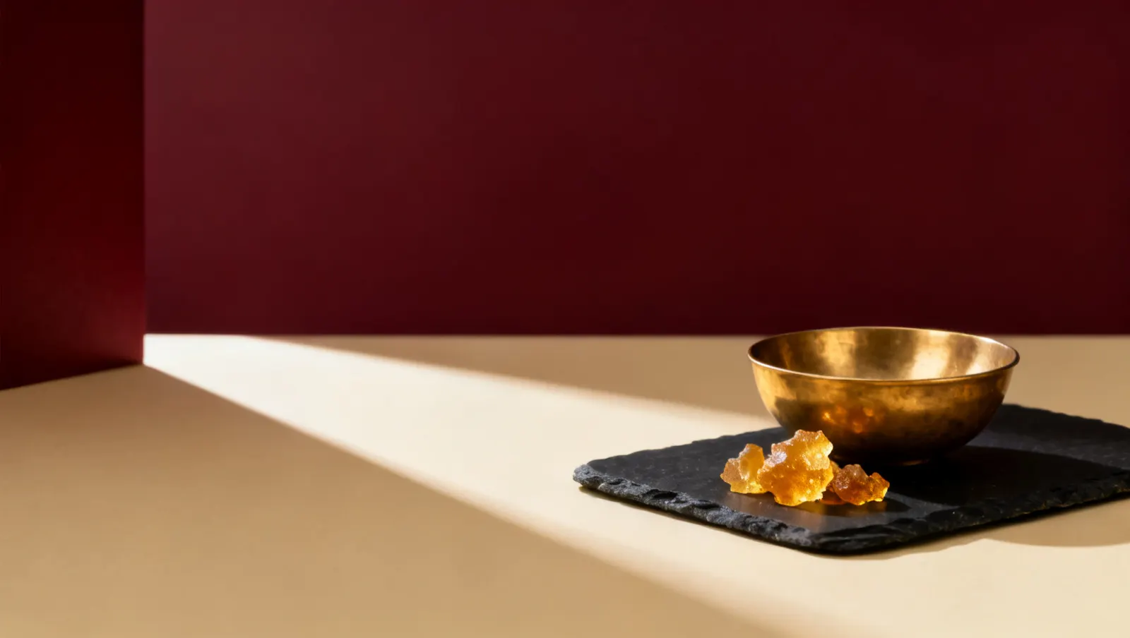 Natureza-morta em luz lateral dura: uma tigela lisa de latao e um bloco de resina de ambar dourado, sobre fundo vinho e creme