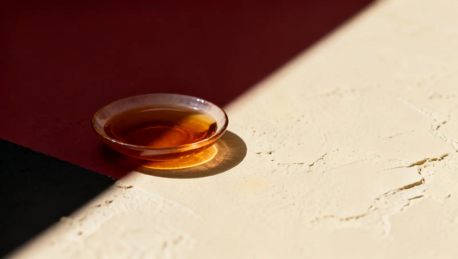 Natureza-morta em luz lateral dura: um prato raso com liquido ambar, sobre fundo vinho e creme