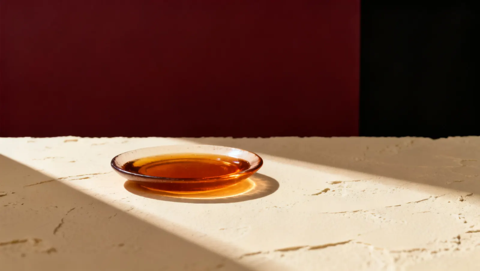 Natureza-morta em luz lateral dura: um prato raso com liquido ambar, sobre fundo vinho e creme