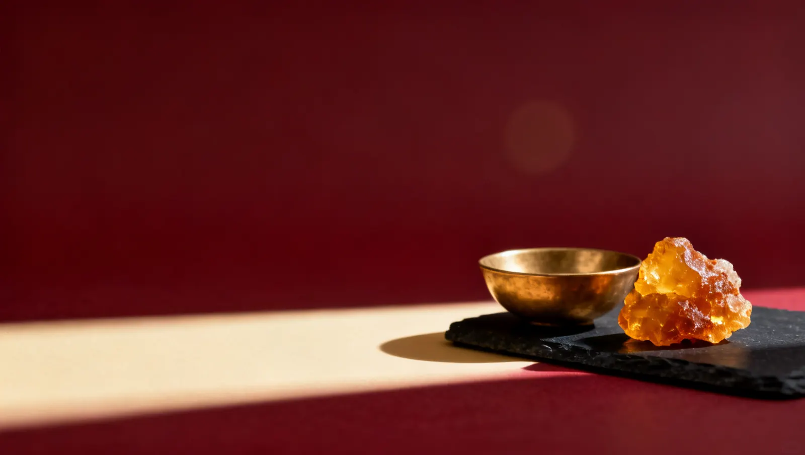 Natureza-morta em luz lateral dura: um bloco de resina de ambar dourado e uma tigela lisa de latao, sobre fundo vinho e creme